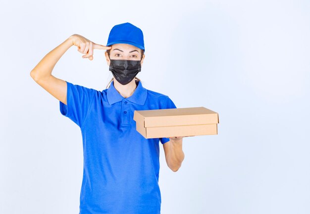 Female courier in blue uniform and face mask holding a cardboard box and thinking about delivery. 