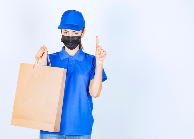 Female courier in blue uniform and face mask holding a cardboard shopping bag and has an idea or thinking. 