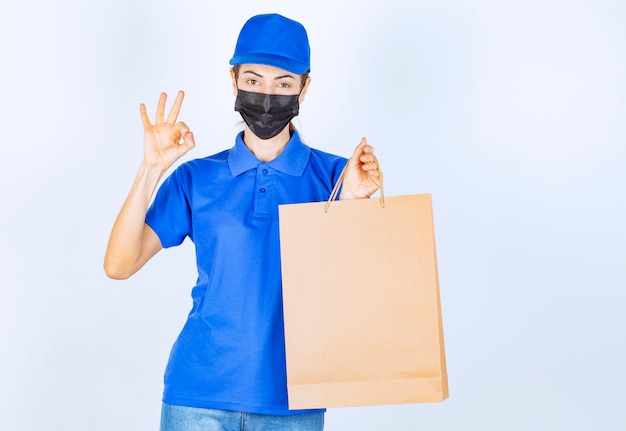 Female courier in blue uniform and face mask holding a cardboard shopping bag and showing satisfaction sign. 