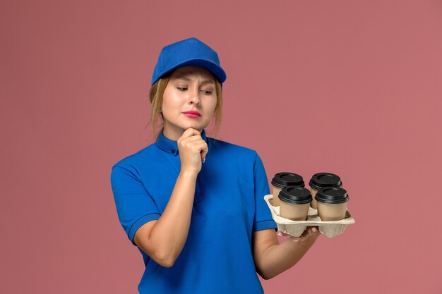 Corriere femminile in uniforme blu che tiene le tazze di consegna marroni di caffè sulla rosa, consegna uniforme dei lavoratori di servizio