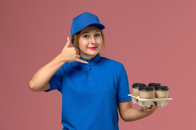 ピンクの笑顔でポーズをとって茶色のコーヒーの配達カップを保持している青い制服の女性の宅配便、サービス制服配達の労働者