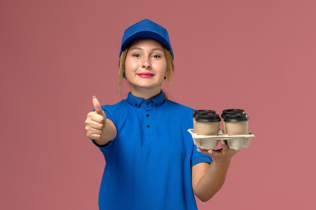 ピンクに笑みを浮かべて茶色のコーヒーの配達カップを保持している青い制服の女性の宅配便、サービス制服配達労働者