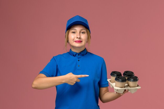 женщина-курьер в синей форме держит коричневые чашки кофе с улыбкой на розовом, доставка униформы работника службы