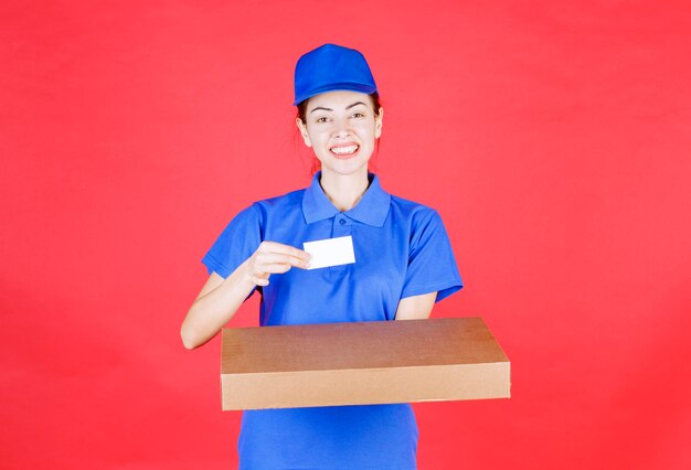 Female courier in blue uniform holding a cardboard box and presenting her business card. 