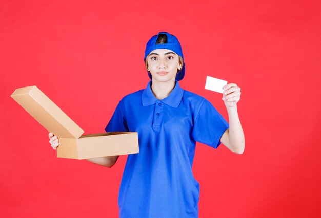 Женский курьер в синей форме держит картонную коробку на вынос и представляет свою визитную карточку.