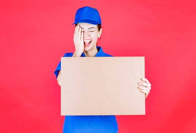 파란색 유니폼을 입은 여성 택배사는 마분지 테이크아웃 피자 상자를 들고 한쪽 눈을 손으로 가리고 있습니다.
