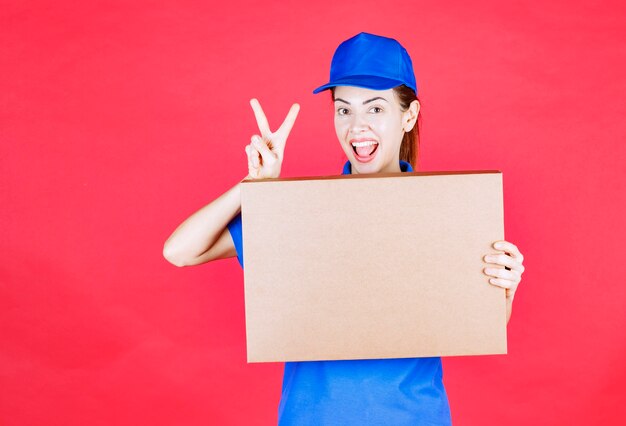 파란색 제복을 입은 여성 택배사는 마분지 테이크아웃 피자 상자를 들고 즐거움 표시를 보여줍니다.