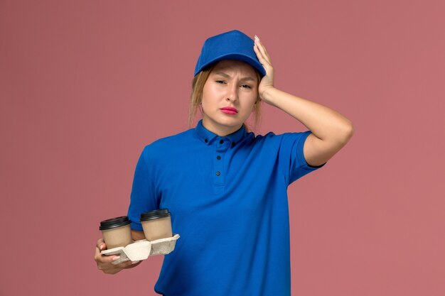 female courier in blue uniform holding cups of coffee having headache on pink, service uniform delivery job