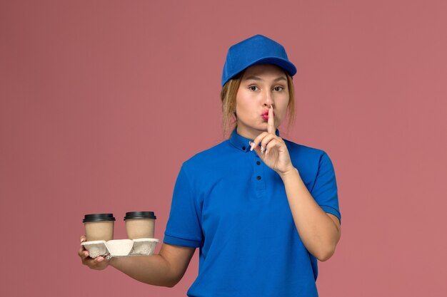 женщина-курьер в синей форме держит чашки кофе, показывая знак тишины на розовом, доставка формы службы