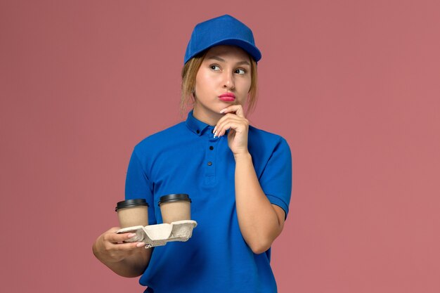 female courier in blue uniform holding cups of coffee thinking on pink, service uniform delivery job