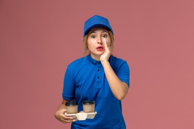 female courier in blue uniform holding cups of coffee whispering on pink, service uniform delivery job