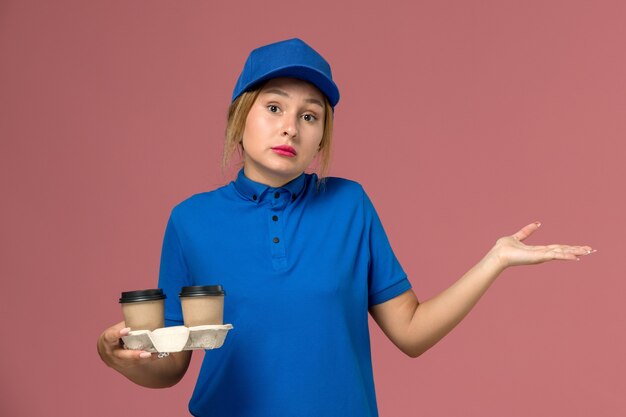 female courier in blue uniform holding cups of coffee with confused expression on pink, service uniform delivery job