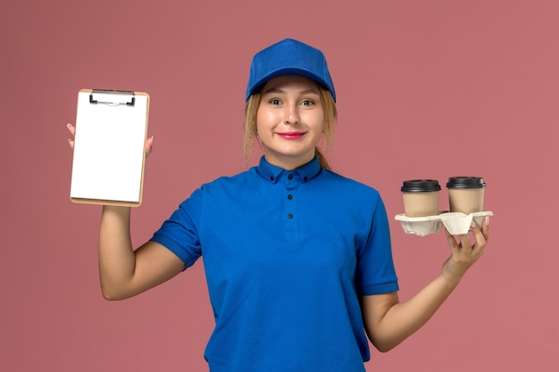 female courier in blue uniform holding delivery cups of coffee and notepad on pink, service job worker uniform delivery