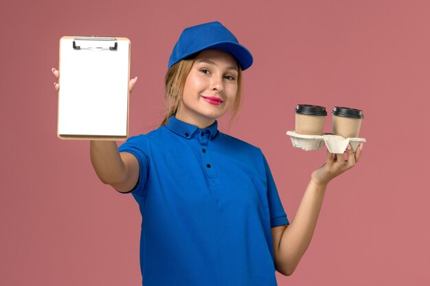 female courier in blue uniform holding delivery cups of coffee and notepad with slight smile on pink, service worker uniform delivery