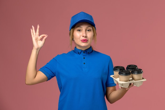женщина-курьер в синей форме с доставкой чашек кофе позирует на розовом, доставка униформы работника службы