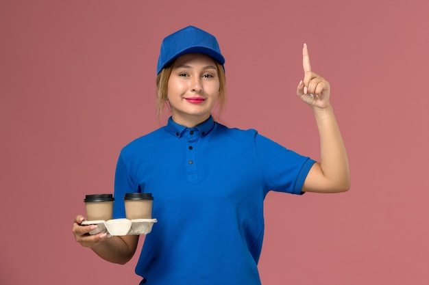 ピンクの上げられた指で微笑んでコーヒーの配達カップを保持している青い制服の女性の宅配便、サービス制服配達の仕事
