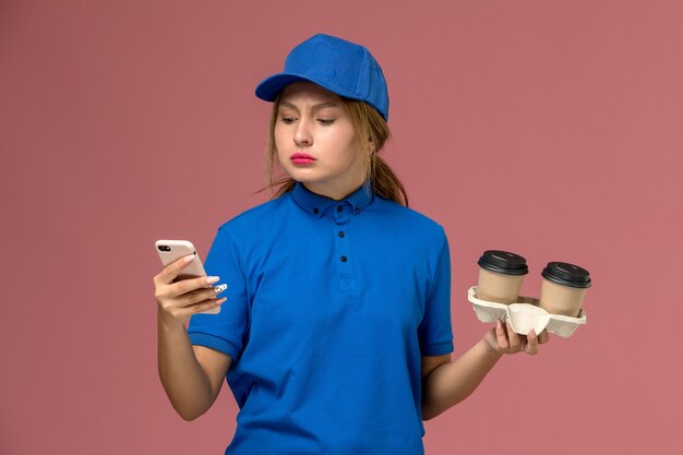 female courier in blue uniform holding delivery cups of coffee and using her phone on pink, service worker uniform delivery