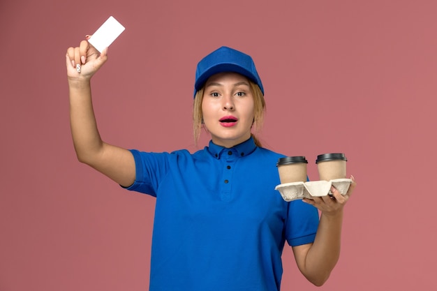 женщина-курьер в синей форме держит чашки с кофе и белую карточку на розовом, работа по доставке служебной формы