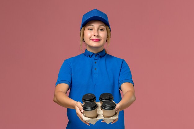 female courier in blue uniform holding delivery cups of coffee with smile on pink, service worker job uniform delivery