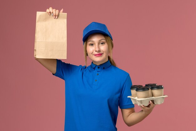 female courier in blue uniform holding food package and brown delivery cups of coffee on light-pink, service uniform delivery job worker