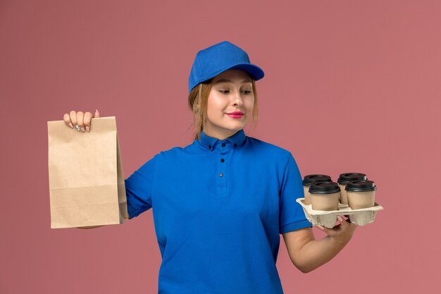 female courier in blue uniform holding food package and brown delivery cups of coffee on pink, service uniform delivery job worker