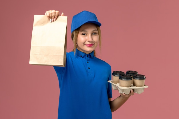 female courier in blue uniform holding food package and brown delivery cups of coffee smiling on pink, service job uniform delivery worker