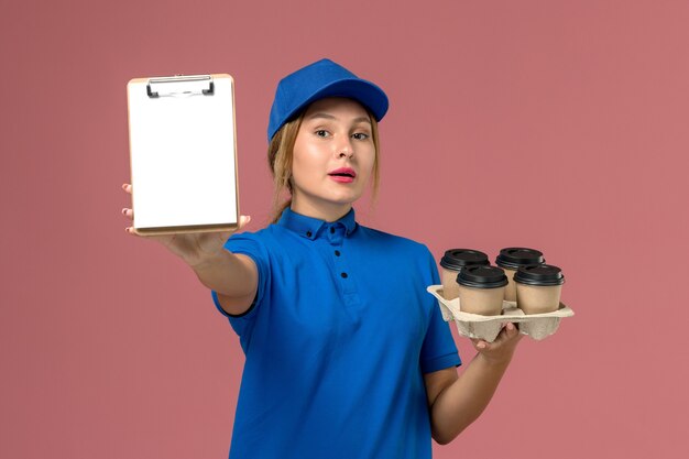 женщина-курьер в синей форме держит блокнот вместе с коричневыми чашками кофе на светло-розовом, доставка рабочей одежды