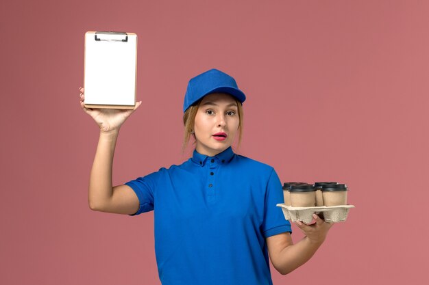 female courier in blue uniform holding notepad along with brown delivery cups of coffee on light pink, job service uniform delivery