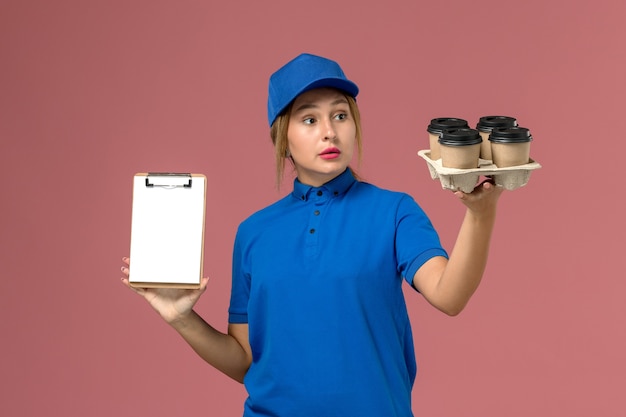 female courier in blue uniform holding notepad along with brown delivery cups of coffee on light-pink, job service uniform delivery