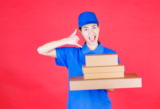 Female courier in blue uniform holding a stock of cardboard boxes and asking for a call. 