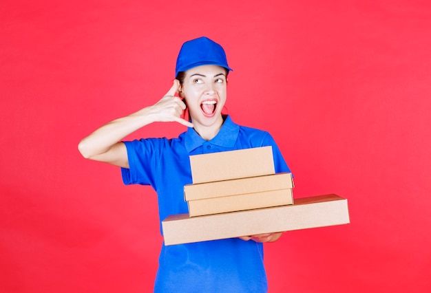 Female courier in blue uniform holding a stock of cardboard boxes and asking for a call. 