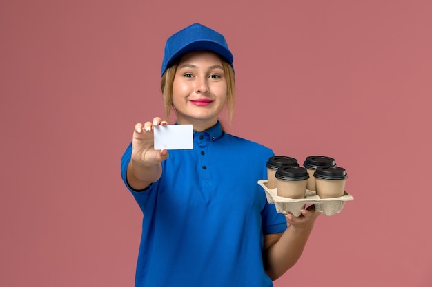 женщина-курьер в синей форме, держащая белую карточку вместе с коричневыми чашками доставки кофе, улыбается на светло-розовом, доставка униформы службы работы