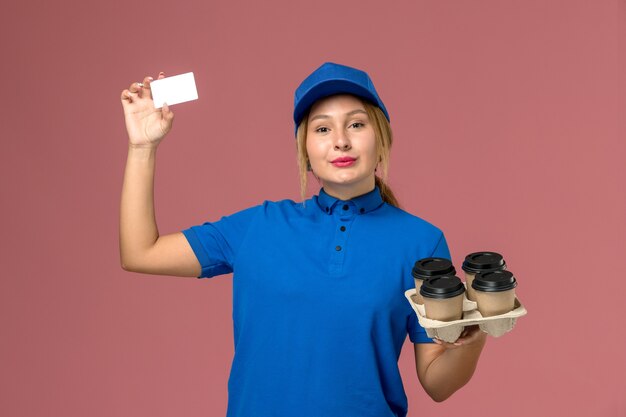 female courier in blue uniform holding white card and brown delivery cups of coffee on pink, service job uniform delivery
