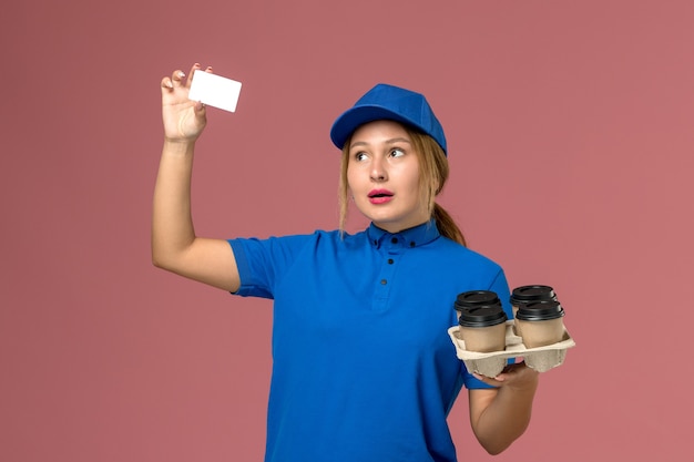 female courier in blue uniform holding white card and brown delivery cups of coffee on pink, service uniform delivery worker