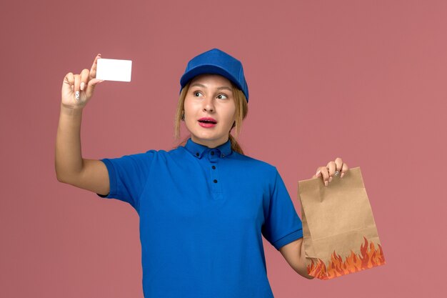 female courier in blue uniform holding white card and food package on light-pink, service uniform delivery job