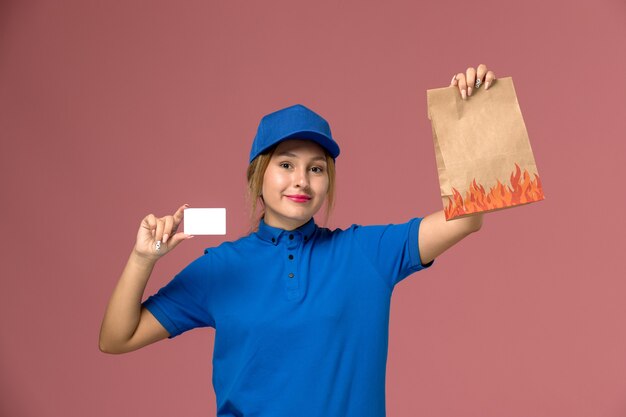 женщина-курьер в синей форме держит белую пластиковую карту и пакет с продуктами на светло-розовом, работа по доставке служебной формы