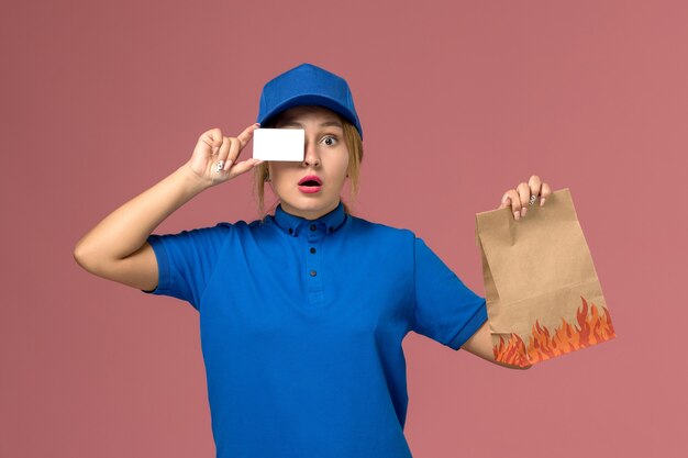 female courier in blue uniform holding white plastic card and food package on light-pink, service uniform delivery