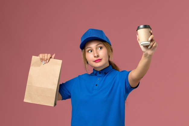 female courier in blue uniform posing holding cup of coffee and food package on pink, service uniform delivery worker