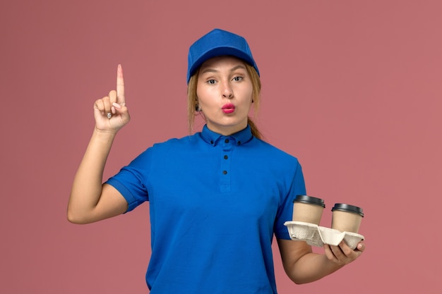 female courier in blue uniform posing holding cups of coffee on pink, service uniform delivery job worker