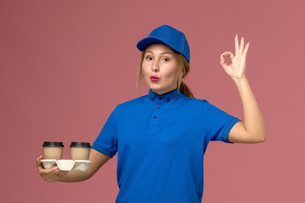 женщина-курьер в синей форме позирует и держит чашки с кофе, показывая знак «хорошо» на розовом, служба доставки униформы