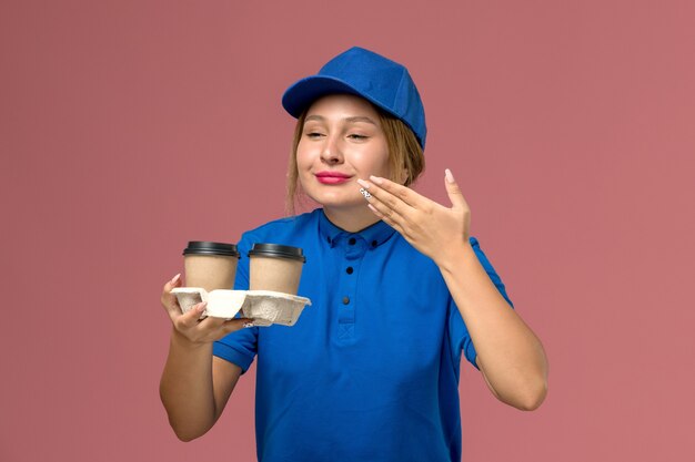 female courier in blue uniform posing and holding cups of coffee smelling on pink, service uniform delivery job