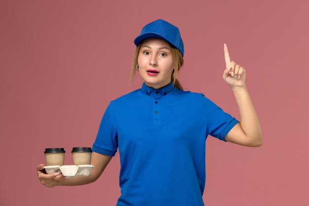 female courier in blue uniform posing and holding cups of coffee with raised finger on pink, service uniform delivery job