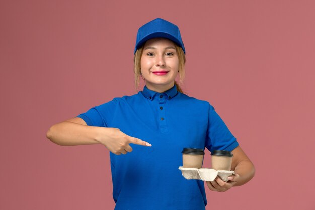 женщина-курьер в синей форме позирует, держа чашки кофе с легкой улыбкой на розовом, работник службы доставки униформы