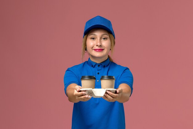 female courier in blue uniform posing and holding cups of coffee with a slight smile on pink, service uniform delivery worker