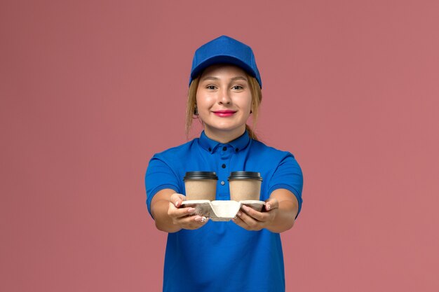 青い制服を着た女性の宅配便がピンク色の微笑みを浮かべてコーヒーのカップをポーズし、保持し、サービス制服配達労働者