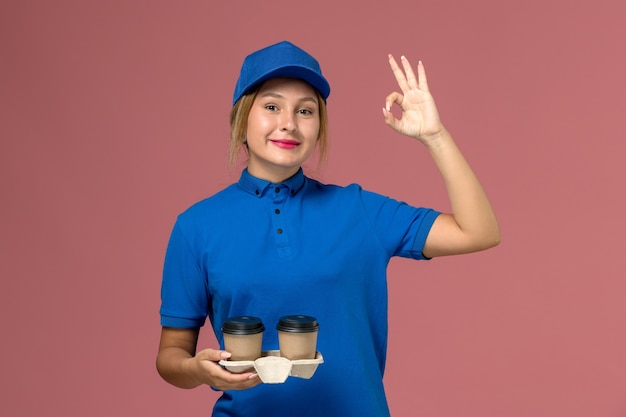 женщина-курьер в синей форме позирует и держит чашки кофе с улыбкой на розовом, служащий службы доставки униформы