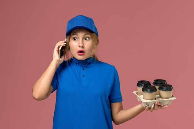 淡いピンクのコーヒーの茶色のカップを保持している電話で話している青い制服の女性の宅配便、サービス制服の配達の仕事