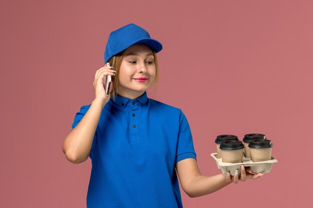 淡いピンクに笑みを浮かべて茶色のコーヒーカップを保持している電話で話している青い制服を着た女性の宅配便、サービス制服配達の仕事