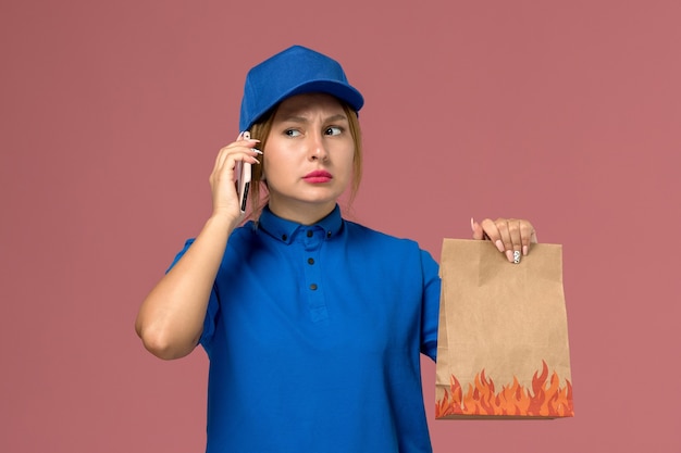 淡いピンクの食品パッケージを保持している電話で話している青い制服の女性の宅配便、サービス制服の配達の仕事