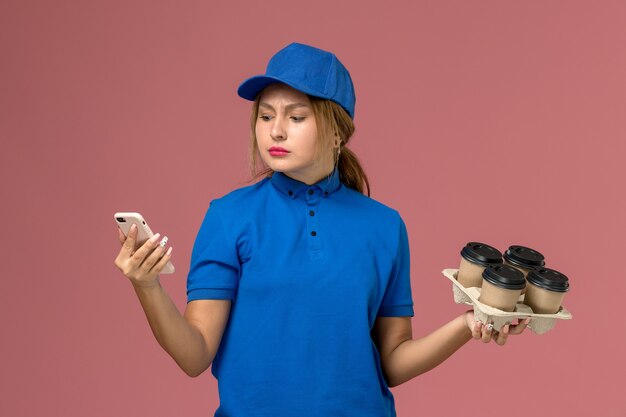 female courier in blue uniform using her phone along with brown cups of coffee on light-pink, work service uniform delivery